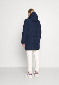 Cappotto puffer blu navy con cappuccio, dotato di vita elasticizzata, cuciture visibili e tasche laterali. Abbinato a pantaloni bianchi e sneaker.