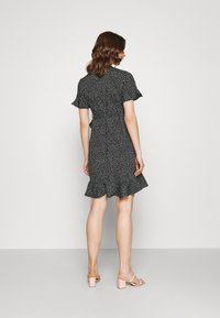 Vestido preto com bolinhas brancas, com mangas curtas e soltas, cintura ajustada e barra ampla. Combinado com sandálias de salto cor-de-rosa claro.