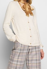 Cremfarbener Cardigan mit Knopfverschluss, zwei Fronttaschen, gerippten Bündchen, kombiniert mit karierten Hosen in Schwarz, Rot und Gelb.