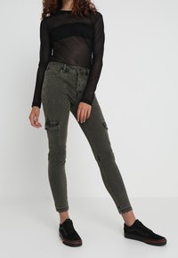 Olivgröna cargo-byxor med slim fit, fickor på sidorna och franskant, tillsammans med en svart genomskinlig långärmad topp och svarta sneakers.