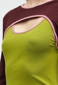 Τοπ μακρυμάνικο με σκοτεινό μπορντό επάνω μέρος και φωτεινό πράσινο κάτω τμήμα. Διαθέτει ροζ αντίθετη ραφή και λεπτομέρεια με κοψίματα στο ντεκολτέ.