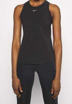 Frau trägt ein ärmelloses schwarzes Nike Tanktop und schwarze Leggings und steht vor einem weißen Hintergrund.