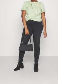 Top corto de color verde claro con dobladillo festoneado, atado en la cintura; combinado con pantalones vaqueros skinny negros y botas negras. Sosteniendo un bolso negro.