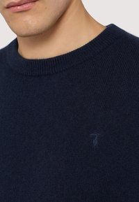 Mörkblå stickad tröja med rund halsringning och ribbad kant, med en subtil märkeslogotyp på bröstet. Mjuk textur.