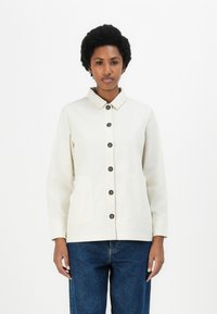 Beige colbert met knopen aan de voorkant, twee zakken en lange mouwen, gemaakt van een gladde stof; gecombineerd met blauwe denim jeans.