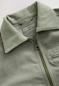 Close-up of light green jacket collar with corduroy texture, partially unzipped brass zipper, and visible inner label marked medium size.
