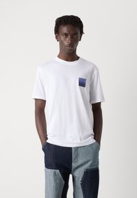 White cotton t-shirt with a square blue pocket, featuring the logo "HUGO." Model wears denim patchwork pants with varying blue tones.