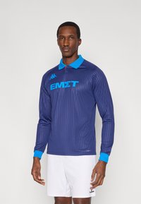 Maglia sportiva a maniche lunghe blu navy con colletto e polsini azzurri. Presenta linee verticali e testo blu. Logo sulla parte superiore sinistra.