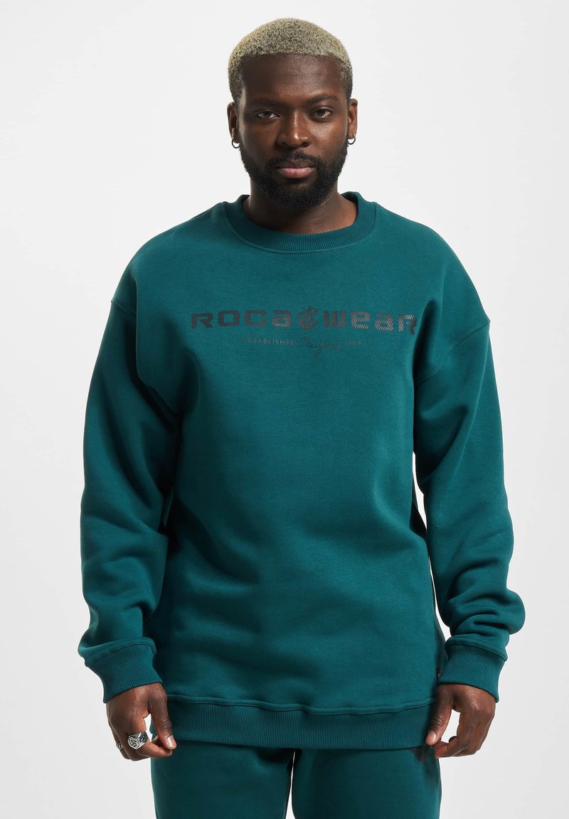 Teal színű pulóverek, textúrált pamut keverékből, bordás mandzsettával és nyakkal. A mellkason fekete "ROCAWEAR" logó található.