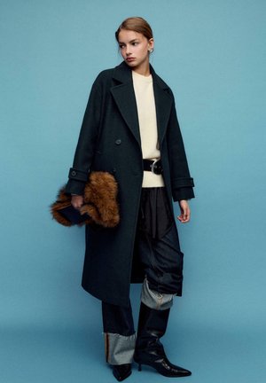 Cappotto lungo verde scuro con grandi risvolti, maglione beige, cintura nera, pantaloni di denim con risvolti, e stivali neri con punte appuntite. Borsa clutch in pelliccia marrone come dettaglio.