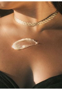 Krämig lotion applicerad på ett solbränt nyckelben, med en flätad metallisk choker vilande på huden. Mjuk textur, mjuk glans.