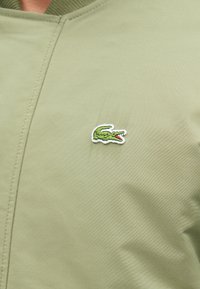 Πράσινο ελαφρύ μπουφάν με λείο υλικό, που διαθέτει ένα μικρό κεντημένο λογότυπο κροκοδείλου σε πράσινο, κόκκινο και λευκό χρώμα στο στήθος.