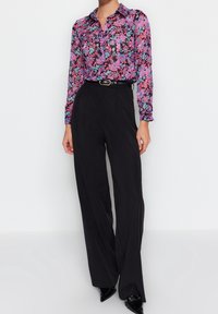 Blusa floral de manga larga, con patrones en rosa, azul y morado, combinada con pantalones negros de tiro alto. Cintura ceñida con un cinturón, zapatos negros elegantes.
