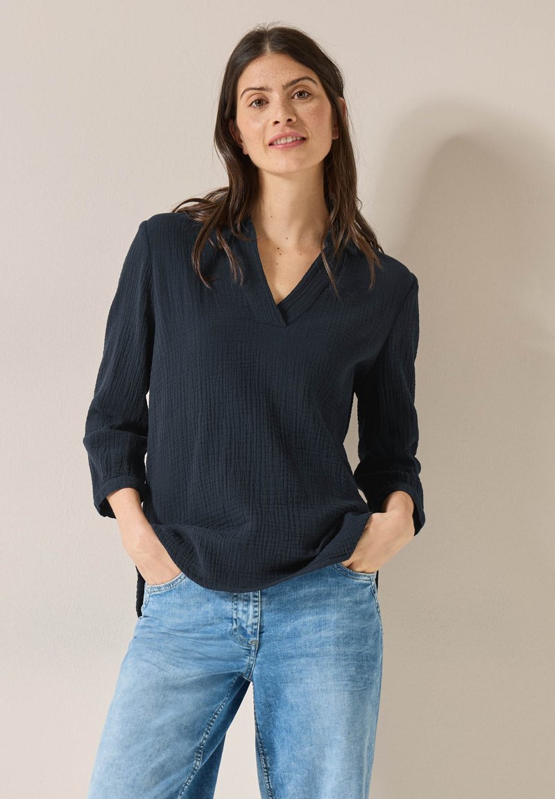 Maglietta a maniche lunghe navy con scollo a V, in tessuto testurizzato e dalla vestibilità rilassata, abbinata a jeans azzurri. Tasche frontali visibili.