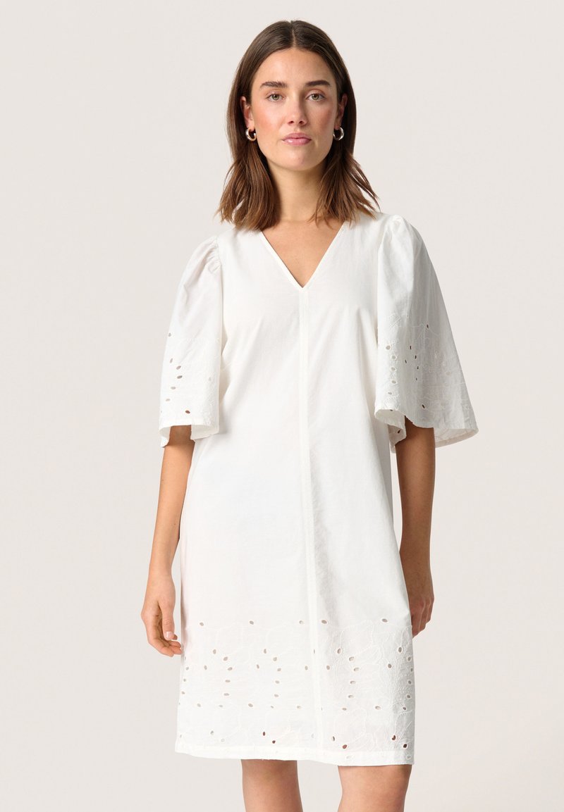 Femme portant une robe blanche longueur genou avec un col en V et des manches mi-longues bouffantes avec des détails de broderie à jours.
