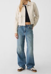 Frau in beigem, gekürztem Jacket über schwarzem Crop-Top, lockeren blauen Jeans und zweifarbigen Sneakers, steht vor weißem Hintergrund.