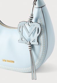 Τσάντα χειρός από δερμάτινο ανοιχτό μπλε, με διακοσμητικό γούρι που απεικονίζει διασταυρούμενα γράμματα «M» και καρδιά, διακοσμημένη με μπεζ ραφές και χρυσό λογότυπο.