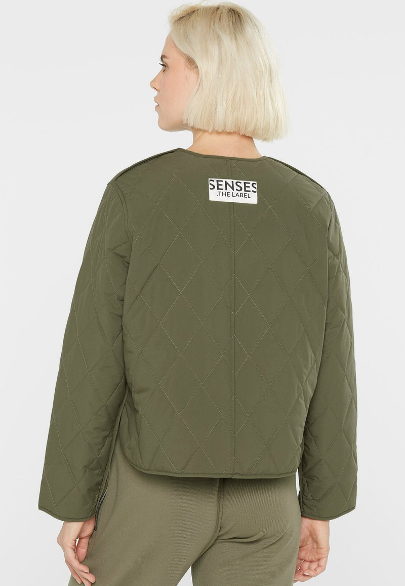 Blonde Person, der eine gesteppte olivgrüne Jacke mit einem "SENSES .THE LABEL"-Aufnäher auf dem oberen Rücken trägt, kombiniert mit passenden Hosen.