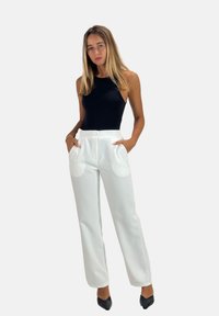 Hvide, højtaljede bukser med sidelommer og knaplukning, kombineret med en sort ærmeløs top, model stående mod en ensfarvet baggrund.