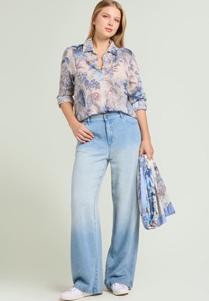 Nainen pukeutuneena puoliläpinäkyvään kukalliseen puseroon ja leveälahkeisiin vaaleansinisiin farkkuihin, pitäen oikeassa kädessään vastaavaa kukkakuviollista huivia.