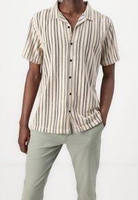 Camicia a maniche corte di colore crema con strisce verticali nere e sottili motivi a pois; presenta un colletto e chiusura con bottoni.