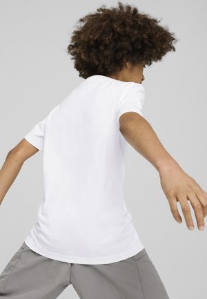 Camiseta blanca de algodón con cuello redondo y mangas cortas, con una textura suave. La persona lleva pantalones cortos grises, vista desde atrás.