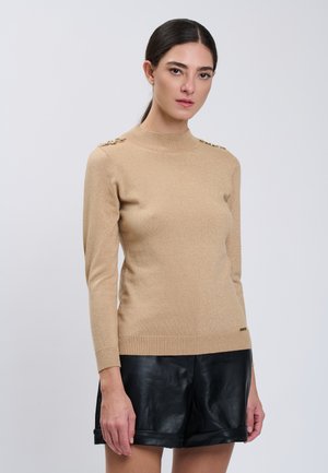 Kvinde med mørkt hår i en beige langærmet sweater med guldknapper på skuldrene og sorte lædershorts, stående mod en ensfarvet grå baggrund.