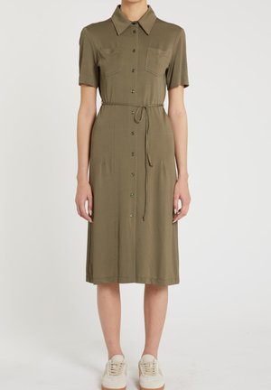 Donna che indossa un vestito camicia verde oliva con bottoni, maniche corte, tasche sul petto, cintura in vita e sneaker bianche, in piedi su sfondo neutro.
