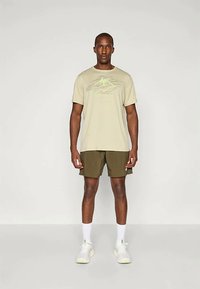 Vaalean beige t-paita, jossa on vihreä graafinen kuvio, yhdistettynä oliivinvihreisiin shortseihin. Kengät ovat valkoiset vaaleilla yksityiskohdilla ja sukat ovat valkoiset.