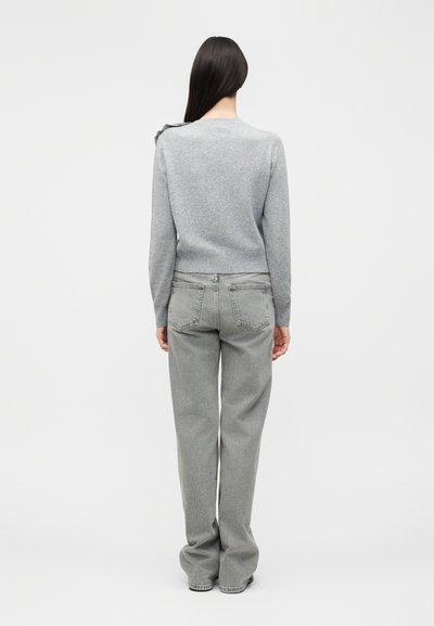 Γυναίκα με μακριά σκουρόχρωμα μαλλιά στέκεται με την πλάτη, φοράει ανοιχτό γκρι πουλόβερ και φαρδιά γκρι τζιν μπροστά σε λευκό φόντο.