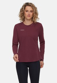 Kvinne med krøllete blondt hår iført en maroon langermet Mammut skjorte og svarte tights, stående mot en hvit bakgrunn.