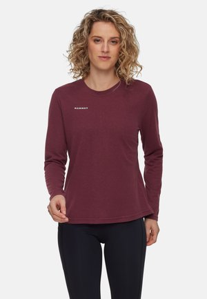 Mujer con cabello rizado rubio que lleva una camiseta de manga larga color burdeos de Mammut y leggins negros, de pie contra un fondo blanco.