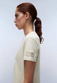 T-shirt in cotone beige con vestibilità classica, scollatura rotonda e maniche corte. Presenta un'applicazione testurizzata cucita sulla manica con il marchio.