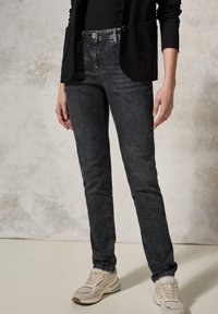 Jeans de denim negro ajustados con una textura ligeramente desvaída, combinados con zapatillas de color claro y una blazer negra.