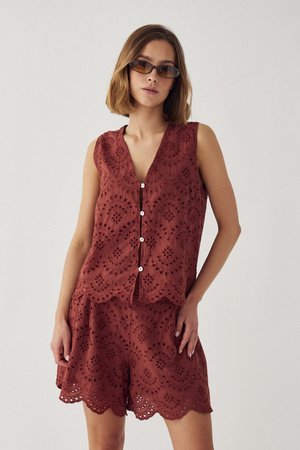 Nő rozsdabarna ujjatlan, gombos felsőt és hozzáillő hímzett rövidnadrágot visel, rövid haja és színezett napszemüvege van, sima fal előtt áll.