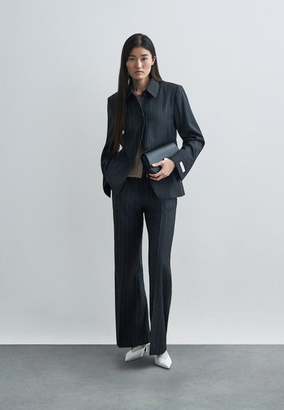 Tumesin triibuline ülikond pikkade varrukatega, laiad püksid ja vormistatud bleiser. Valged terava ninaga kontsad ja must klaht lõpetavad välimuse.