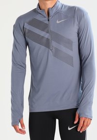 Långärmad, zip-kragnärmst skjorta i ljusgrått med ett strukturerat randmönster. Gjord av elastiskt material, med en reflexlogotyp på bröstet.