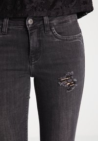Pantalones vaqueros de denim negro con un diseño ajustado, que presentan un detalle desgastado en el muslo y un ligero desvanecimiento. La textura es suave con un estilo clásico de cinco bolsillos.