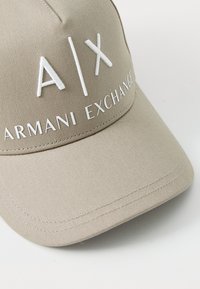 Bežinis beisbolo kepuraitė su baltu siuvinėtu logotipu "A|X" ir "ARMANI EXCHANGE." Pagaminta iš tekstūrinio audinio, turi lenktą kraštą ir siuvinėjimo detales.