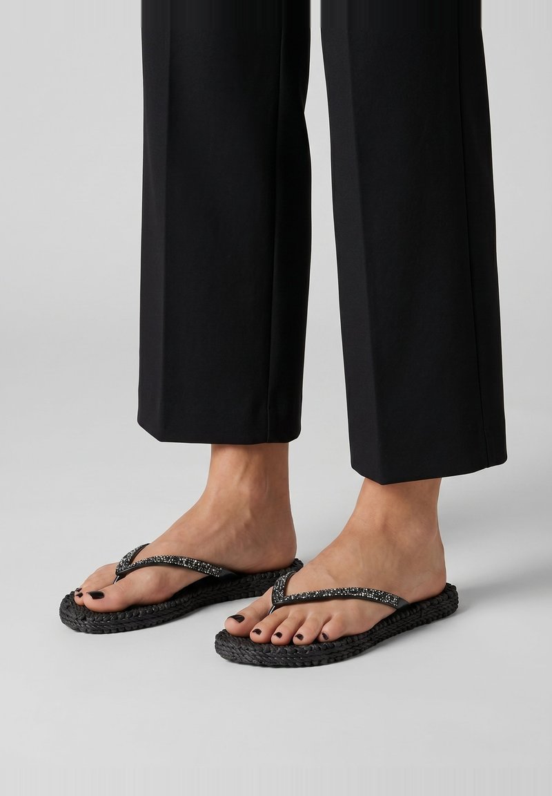 Feet wearing black braided flip-flops with sparkly straps, paired with black cropped pants on a light gray background.