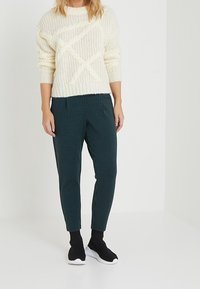 Suéter de punto crema con un patrón texturizado, combinado con pantalones cuadros en verde oscuro. El modelo lleva calcetines negros con suelas blancas.