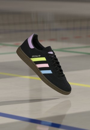Baskets Adidas Spezial noires avec des rayures jaune pastel, rose et bleue, languette de talon violette et semelle marron, suspendues au-dessus du sol d’un terrain intérieur.