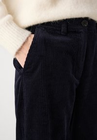 Persoon met een crème trui en hand in de zak van donkere marineblauwe corduroy broek, die de gestructureerde stof en knopen toont.