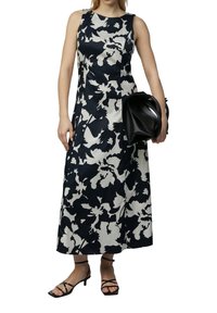 Maxikleid mit floralem Muster in Marineblau und Weiß, ärmelloses Design, gerader Schnitt, kombiniert mit einer schwarzen Handtasche und Sandalen mit Riemchen.