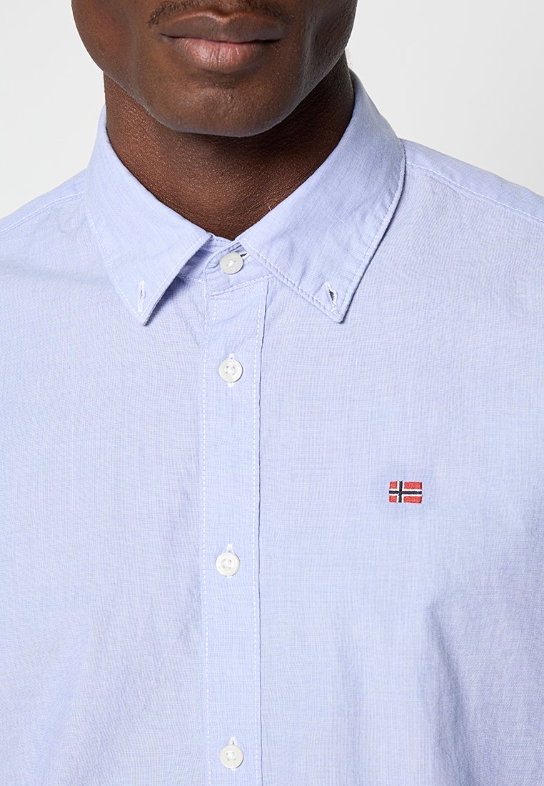 Camicia azzurra chiara con tessuto testurizzato, con una piccola bandiera norvegese ricamata sul petto e bottoni bianchi standard.