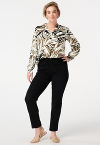 Femme debout avec la main sur la hanche portant un chemisier à motifs de feuilles marron, noir et blanc, un pantalon noir et des chaussures à talon compensé tissées beiges.