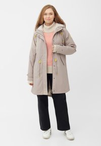 Lichtgrijze parka met een fuzzy capuchonvoering, gele accenten en een rits aan de voorkant. Draagt over een crèmekleurige trui met rode strepen en zwarte broek.