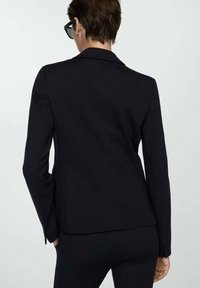 Blazer nero su misura con un design aderente, texture liscia, chiusura a un bottone e dettagli di cucitura discreti sulla parte posteriore.