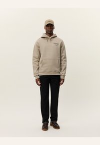 Sudadera beis con bolsillo frontal, capucha con cordón y detalle del logo; pantalones negros; zapatos marrones; gorra beige. Textura de tela suave.