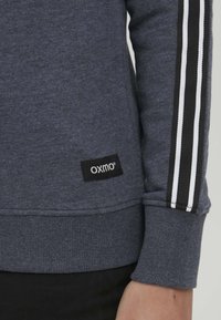 Felpa grigia scura con dettagli a righe bianche e nere sulle maniche, caratterizzata da un polsino a costine e un patch con il logo "OXMO".